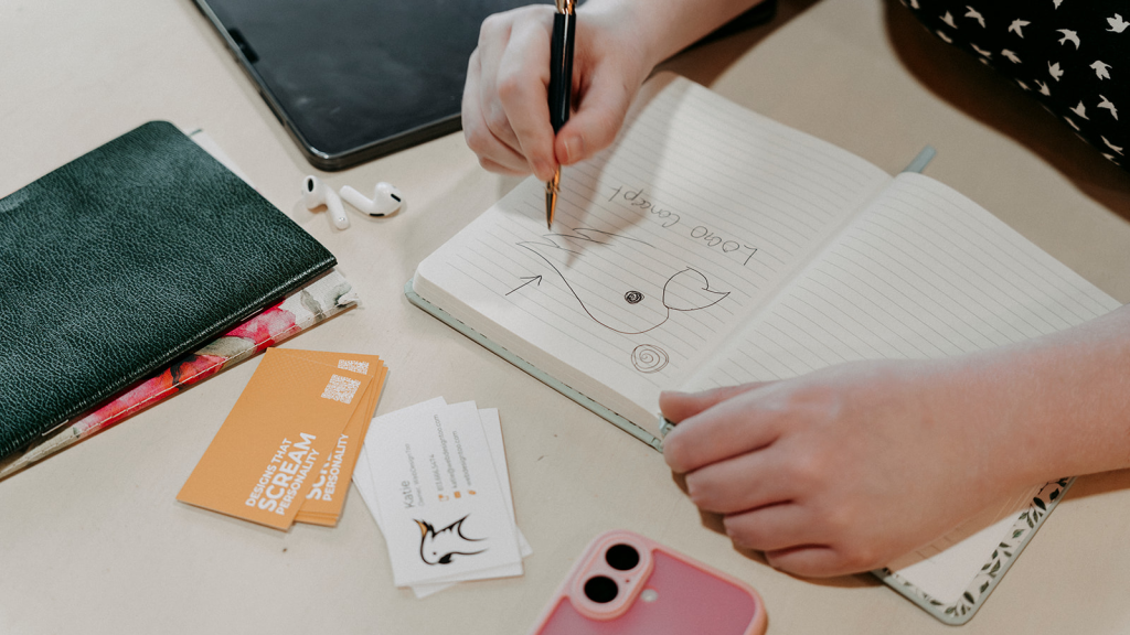 Woman drawing a preliminary sketch of a cockatoo logo, with business cards and notebooks around her