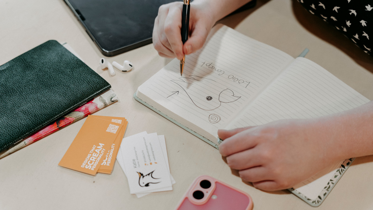 Woman drawing a preliminary sketch of a cockatoo logo, with business cards and notebooks around her
